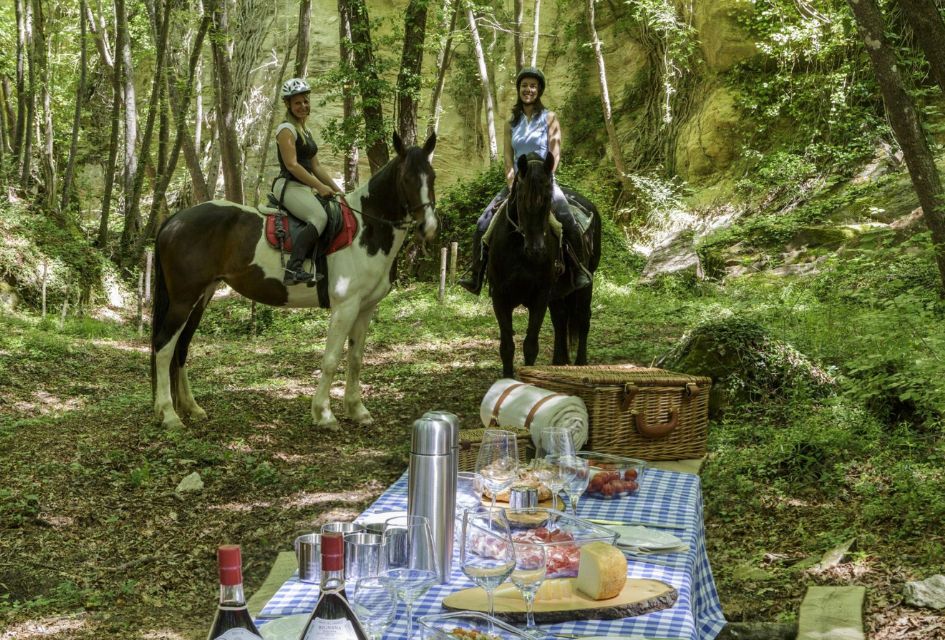 Horse-riding at Borgo Pignano Estate Two women on horses standing in a forest behind a picnic table with charcuterie and wine, a great group activity for your Tuscany holiday.