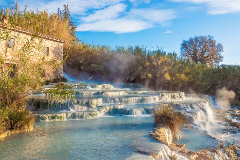 The Saturnia hot springs, a fantastic activity to do while enjoying your luxury holidays in Tuscany. Blue water ripples over multiple layers, with a house to one side and blue skies above.