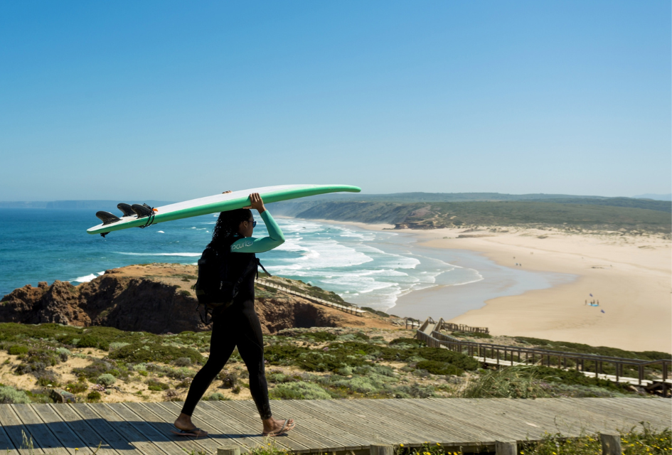 Surf Beaches in the Algarve (Credit ©ATAlgarve) Female surfer carrying a soft surf board down a wooden walkway towards one of the top surf beaches in the Algarve