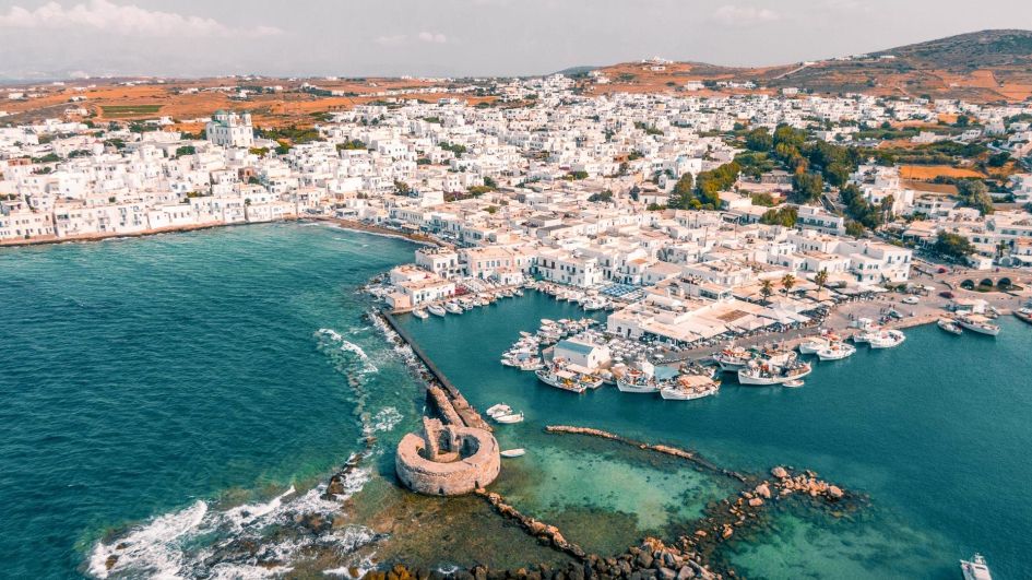 An aerial shot of a harbour in Paros depicting the classic white Cycladic architecture of its buildings and rolling hills in the background.