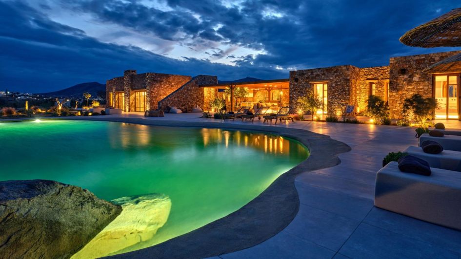 Villa Desert Rose at dusk. The enchanting stone building is lit up in the background, whilst the foreground shows an illuminated pool - perfect for relaxing on luxury holidays in Paros.