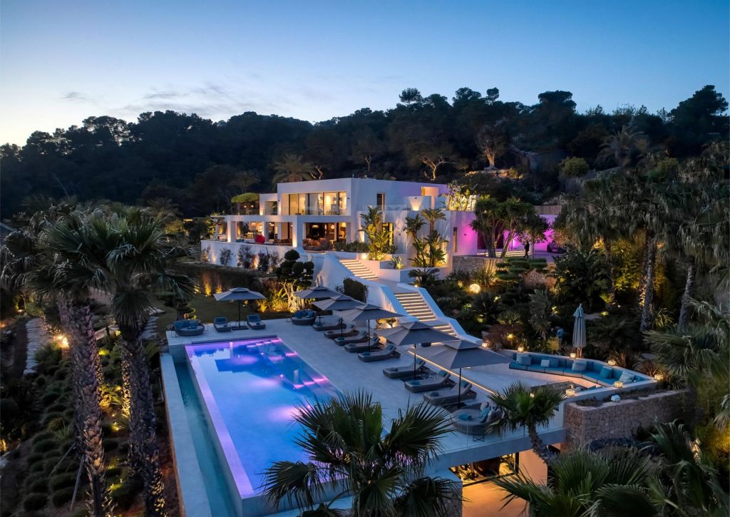 Villa de Sueños, Ibiza View of luxury Villa de Sueños in Ibiza at dusk, with lights illuminating the gardens and surrounding woodland