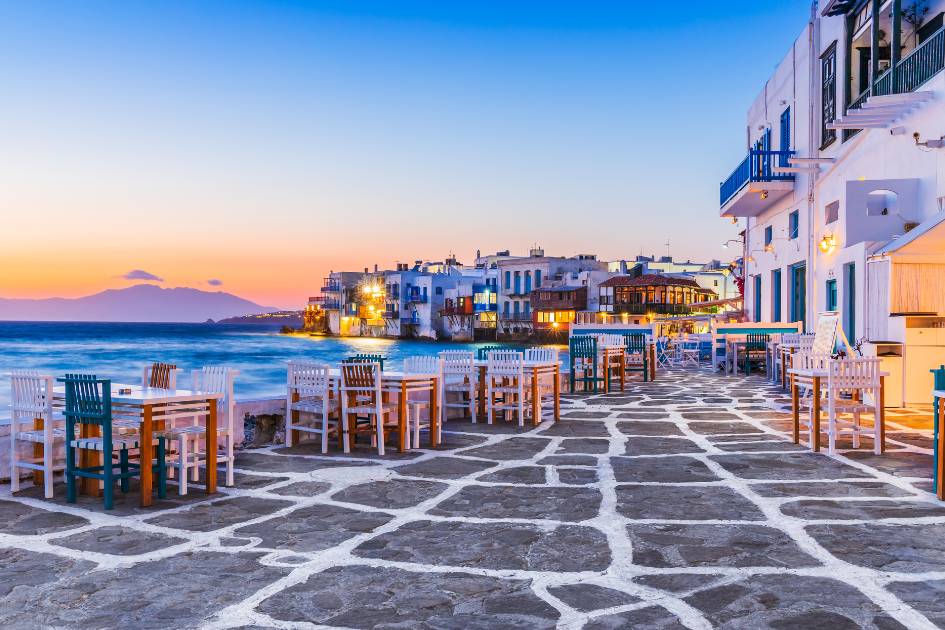 Waterfront bar at sunset in Mykonos,. Greece. Larg cobbled stones, [ink hues, mountain and coean view, and sparkling lights by the water.