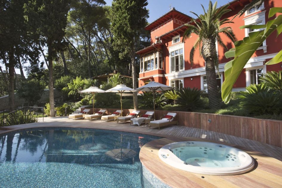 The luscious private outdoor space at Captain's Villa Rouge, featuring a swimming pool, hot tub, loungers, terrace and towering trees, before the villa's distinctive red exterior.