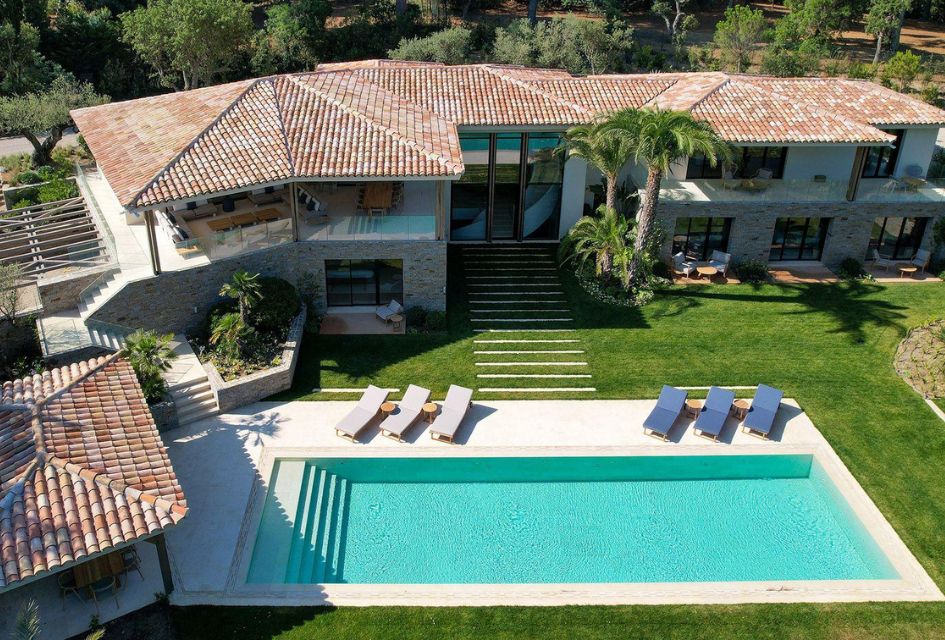 Exterior of Villa B in St Tropez, with a large outdoor pool and sun loungers, and a wraparound building with palm trees.