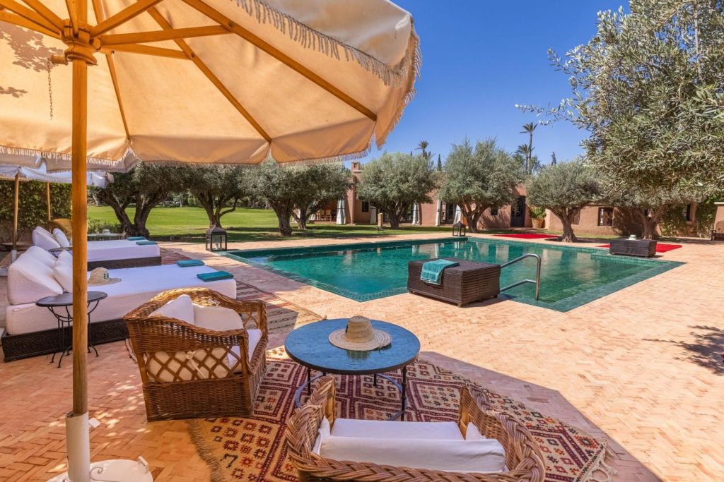 Ezzahra Estate, Marrakech Across 5 villas, expansive gardens and beautiful wellness inside and out, Ezzahra Estate shines as one of the best wellness villas around the world! This image of one of its outdoor pools, surrounded by loungers and vegetation, depicts a scene that's made for relaxation under clear blue skies.